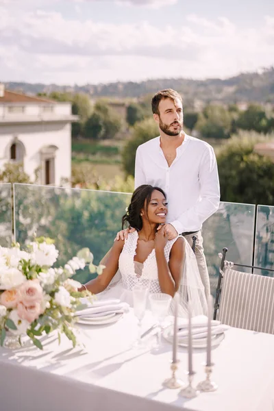 Destination fine-art γάμος στη Φλωρεντία, Ιταλία. Αφροαμερικανή νύφη κάθεται σε ένα τραπέζι του γάμου, Καυκάσιος γαμπρός αγκαλιάζει τους ώμους της. Πολυφυλετικό ζευγάρι γάμου — Φωτογραφία Αρχείου