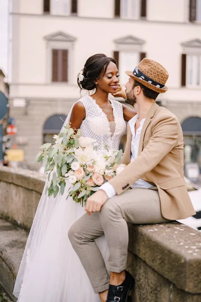 Γάμος στη Φλωρεντία, Ιταλία. Διαφυλετικό ζευγάρι γάμων. Αφροαμερικανή νύφη με λευκό φόρεμα με μακρύ πέπλο και μπουκέτο, και Καυκάσιος γαμπρός με αμμώδες σακάκι και ψάθινο καπέλο. — Φωτογραφία Αρχείου