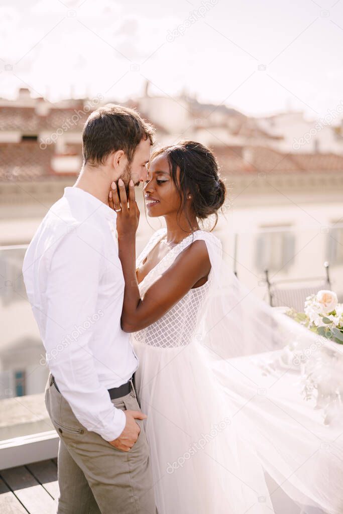 Mixed-race wedding couple. Caucasian groom and African-American bride cuddling on a rooftop in sunset sunlight. Destination fine-art wedding in Florence, Italy. 