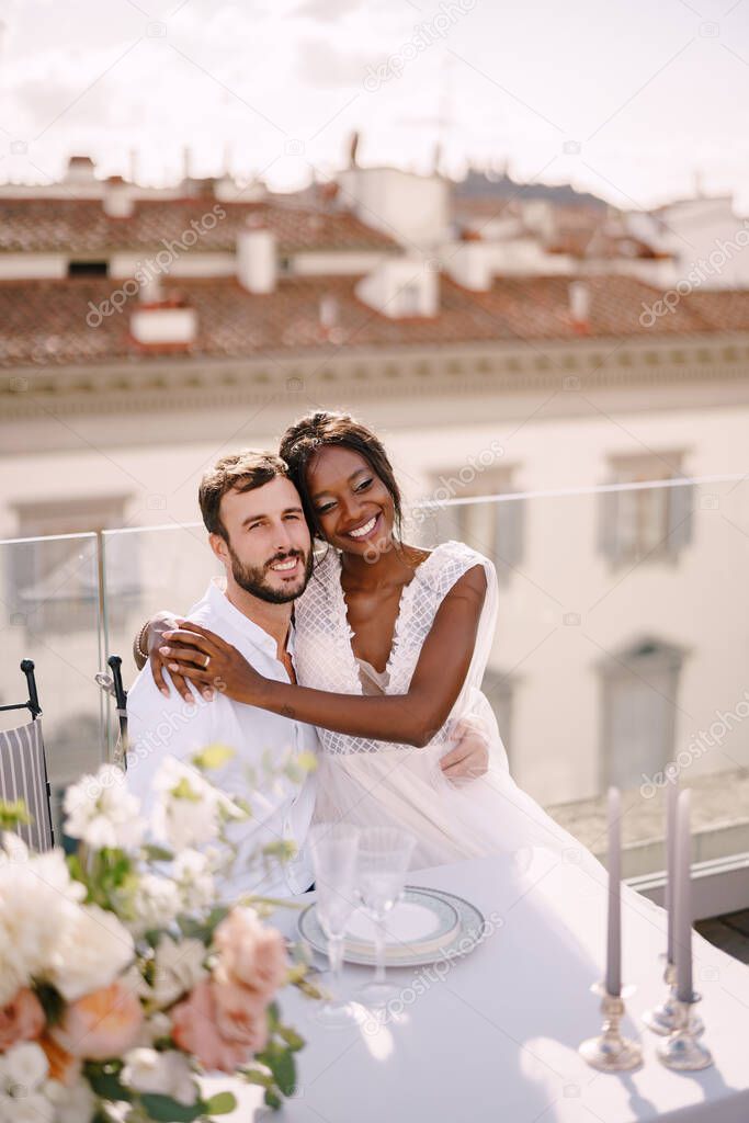 Interracial wedding couple. Destination fine-art wedding in Florence, Italy. African-American bride and Caucasian groom are sitting at the rooftop wedding dinner table overlooking the city.