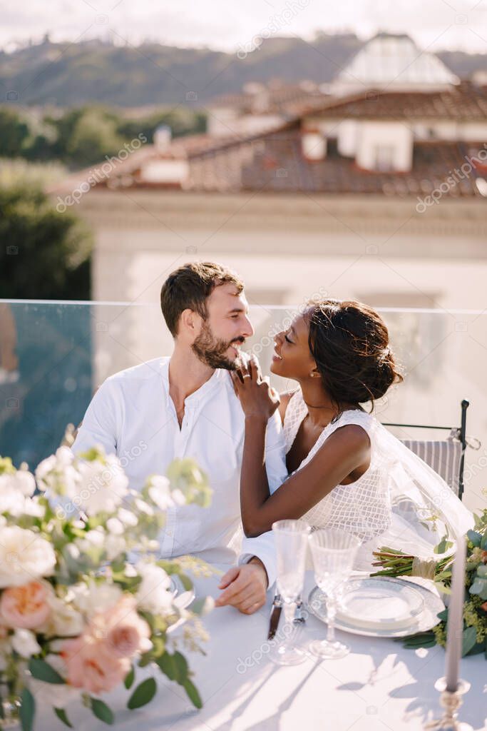 Destination fine-art wedding in Florence, Italy. Multiethnic wedding couple. African-American bride and Caucasian groom are sitting at the rooftop wedding dinner table overlooking the city.