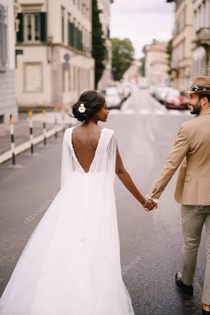 Multiracial wedding couple. Wedding in Florence, Italy. African-American bride in a white dress and Caucasian groom in a hat are walking along the road among cars.
