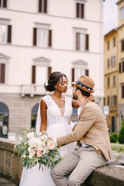 Multi-etnisch bruidspaar. Bruiloft in Florence, Italië. Afro-Amerikaanse bruid in een witte jurk met een lange sluier en een boeket, en Kaukasische bruidegom in een zandjas en strohoed. — Stockfoto