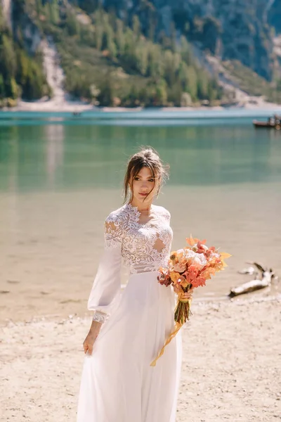 Vacker brud i en vit klänning med ärmar och spets, med en gul höstbukett av torkade blommor och pion rosor, på Lago di Braies i Italien. Destination bröllop i Europa, på Braies sjön. — Stockfoto