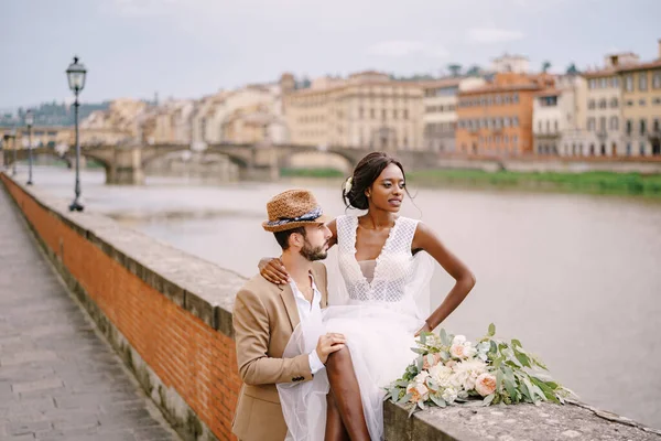 Wedding in Florence, Italy. Multiethnic wedding couple. An African-American bride is sitting on a brick wall and Caucasian groom is hugging her. Arno River Embankment, overlooking city and bridges