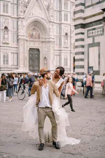 Pasangan pernikahan multietnis. Pernikahan di Florence, Italia. Seorang pengantin Afrika-Amerika melompat ke bahu pengantin pria Kaukasia di Piazza del Duomo. Stok Lukisan  