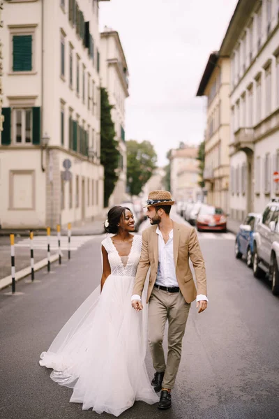Pernikahan di Florence, Italia. Pasangan pernikahan multietnis. Pengantin Afrika-Amerika dengan gaun putih dan pengantin pria Kaukasia dengan topi berjalan di sepanjang jalan di antara mobil. Stok Foto