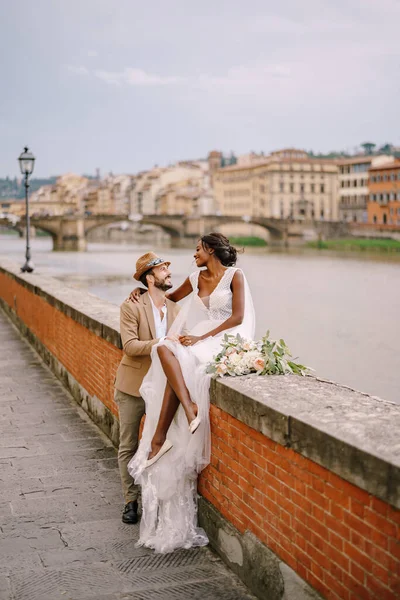 Pasangan pernikahan multirasial. Pernikahan di Florence, Italia. Seorang pengantin wanita Afrika-Amerika duduk di dinding bata dan pengantin pria Kaukasia memeluknya. Arno River Embankment, menghadap kota dan jembatan Stok Gambar