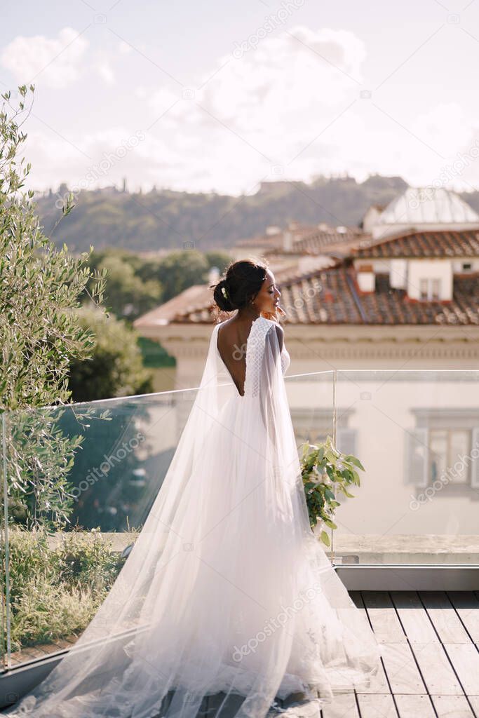 Destination fine-art wedding in Florence, Italy. African-American bride, with a bouquet in her hands, stands on the roof in the sunset light.