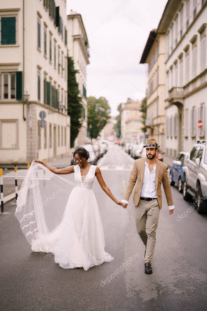 Mixed-race wedding couple. Wedding in Florence, Italy. African-American bride in a white dress and Caucasian groom in a hat are walking along the road among cars.