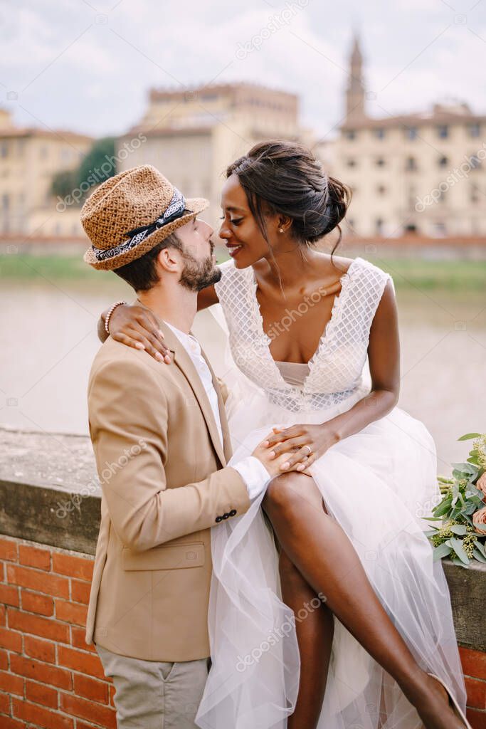 Wedding in Florence, Italy. Interracial wedding couple. An African-American bride is sitting on a brick wall and Caucasian groom is hugging her. Arno River Embankment, overlooking city and bridges