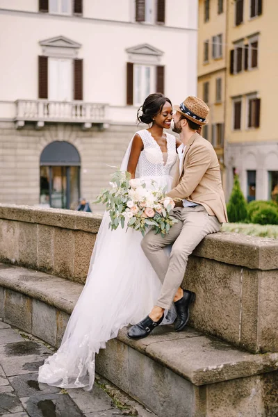 Een gemengd echtpaar. Bruiloft in Florence, Italië. Afro-Amerikaanse bruid in een witte jurk met een lange sluier en een boeket, en Kaukasische bruidegom in een zandjas en strohoed. — Stockfoto