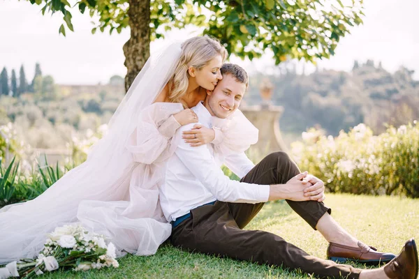 Pernikahan di Florence, Italia, di sebuah villa-winery tua. Pasangan pengantin sedang duduk di atas rumput di taman di bawah pohon, pengantin wanita memeluk pengantin pria. — Stok Foto