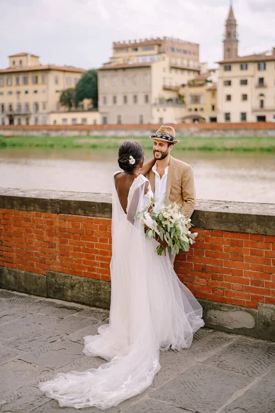 Fajok közötti házaspár. Esküvő Firenzében, Olaszországban. Afrikai-amerikai menyasszony és fehér vőlegény áll ölelés a töltés az Arno folyó, kilátással a városra és a hidak. — Stock Fotó
