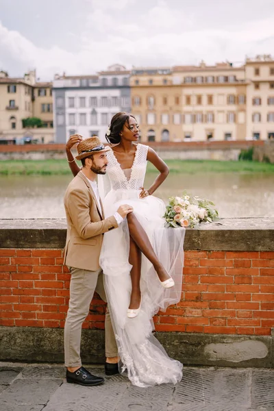 Pasangan pernikahan multietnis. Pernikahan di Florence, Italia. Seorang pengantin wanita Afrika-Amerika duduk di dinding bata dan pengantin pria Kaukasia memeluknya. Arno River Embankment, menghadap kota dan jembatan — Stok Foto