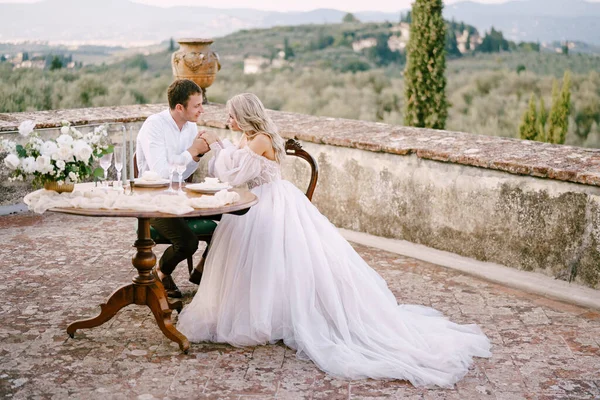 Pernikahan di sebuah vila anggur tua di Tuscany, Italia. Pasangan pengantin sedang duduk di meja makan di atap sebuah villa tua, pengantin pria memegang pengantin wanita dengan tangan. — Stok Foto