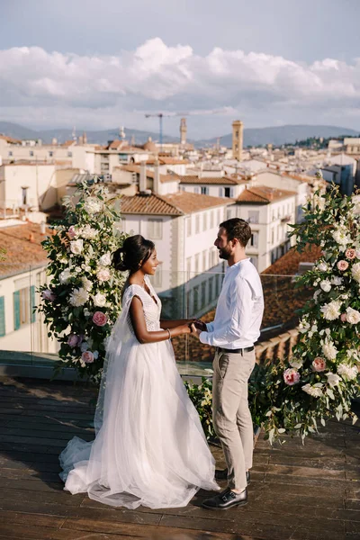 Destination fine-art wedding in Florence, Italy. A wedding ceremony on the roof of the building, with cityscape views of the city and the Cathedral of Santa Maria Del Fiore. Multiracial wedding couple