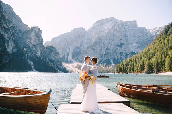 Das Brautpaar spaziert an einem hölzernen Bootsanleger am Pragser See in Italien entlang. Hochzeit in Europa, am Pragser See. Frischvermählte spazieren, küssen, umarmen sich vor dem Hintergrund felsiger Berge. — Stockfoto