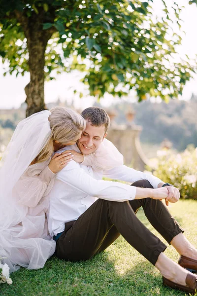 Pernikahan di Florence, Italia, di sebuah villa-winery tua. Pasangan pengantin sedang duduk di atas rumput di taman di bawah pohon, pengantin wanita memeluk pengantin pria. — Stok Foto
