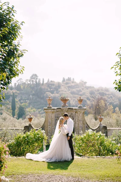 The bride and groom walk in the park. Wedding at an old winery villa in Tuscany, Italy. Round wedding arch decorated with white flowers and greenery in front of an ancient Italian architecture. — Stock Photo, Image