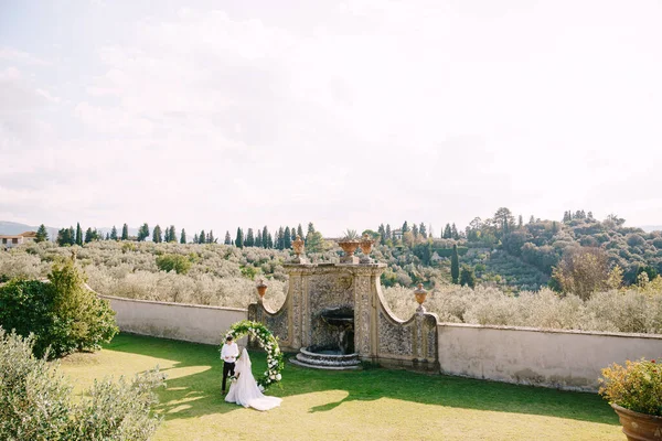 Wedding at an old winery villa in Tuscany, Italy. Wedding couple under a round arch of flowers. The groom reads wedding vows. — Stock Photo, Image