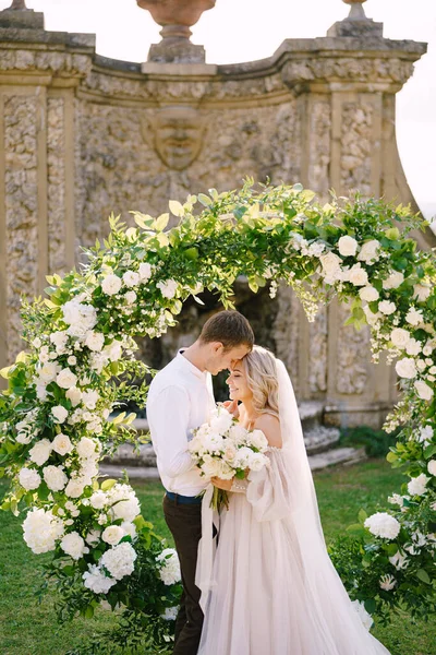 Pernikahan di sebuah vila anggur tua di Tuscany, Italia. Pernikahan pasangan dekat lengkungan pernikahan bulat dihiasi dengan bunga putih dan hijau di depan arsitektur Italia kuno. — Stok Foto