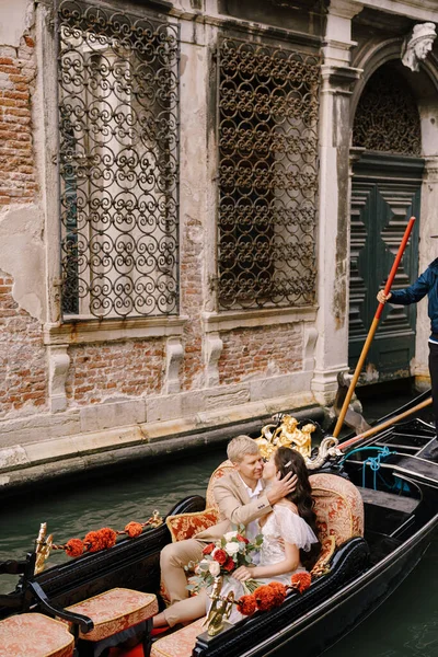 Italia boda en Venecia. Gondolier rueda novia y novio en una clásica góndola de madera a lo largo del estrecho canal veneciano. Los recién casados están sentados en barco nariz a nariz, nadan contra el telón de fondo de celosía forjada — Foto de Stock