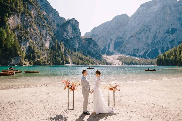 Pengantin pria mengenakan cincin untuk pengantin wanita, di tempat untuk upacara, dengan lengkungan kolom bunga musim gugur, dengan latar belakang Lago di Braies di Italia. Tujuan pernikahan di Eropa, di danau Braies Stok Foto Bebas Royalti