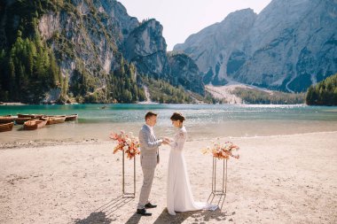 Gelin, İtalya 'daki Lago di Braies' in arka planına karşı, sonbahar çiçek sütunlarından bir kemerle tören için damada yüzük takar. Düğün Avrupa 'da, Braies Gölü' nde..