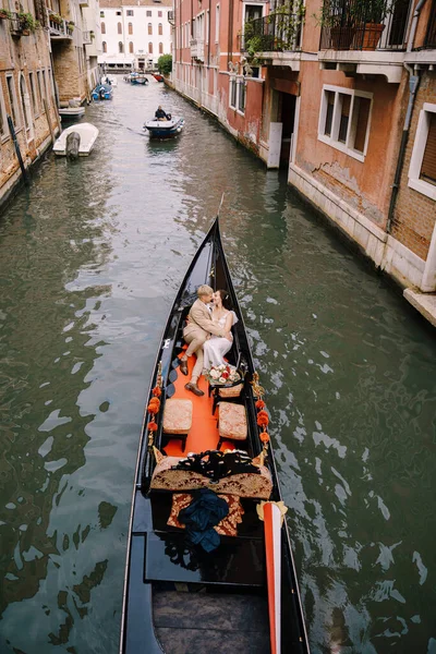 Італійське весілля у Венеції. Гондольєр кидає наречену з нареченим у класичну дерев'яну гондолу по вузькому венеціанському каналу. Молоді люди сидять у човні на задньому плані стародавніх будинків.. — стокове фото