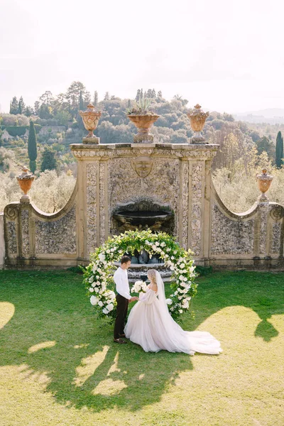 Pernikahan di sebuah vila anggur tua di Tuscany, Italia. Pernikahan pasangan dekat lengkungan pernikahan bulat dihiasi dengan bunga putih dan hijau di depan arsitektur Italia kuno. — Stok Foto