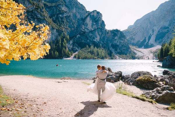 Mariée et marié sous un arbre d'automne, au Lago di Braies en Italie. Mariage de destination en Europe, sur le lac de Braies. Les jeunes mariés amoureux dansent et tournent . — Photo