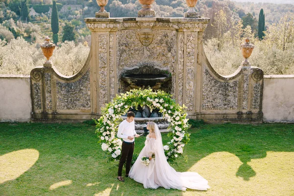 Casamento em uma antiga vinícola villa na Toscana, Itália. Casamento par abaixo de um arco redondo de flores. O noivo lê votos de casamento . — Fotografia de Stock