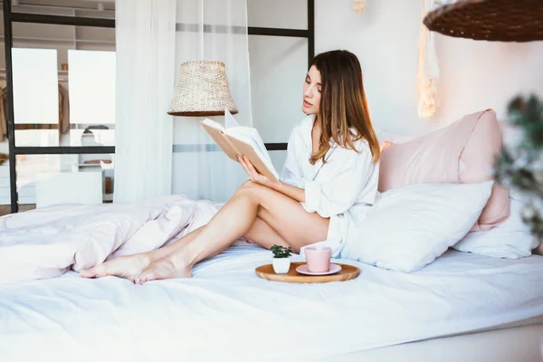 Fille lisant un livre, buvant du café ou du thé, se reposant au lit . — Photo