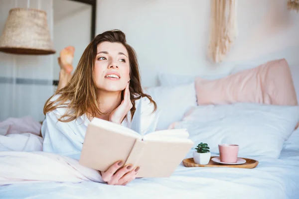 Fille lisant un livre, buvant du café ou du thé, se reposant au lit . — Photo