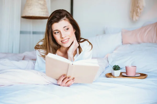 Chica leyendo un libro, tomando café o té, descansando en la cama . — Foto de Stock