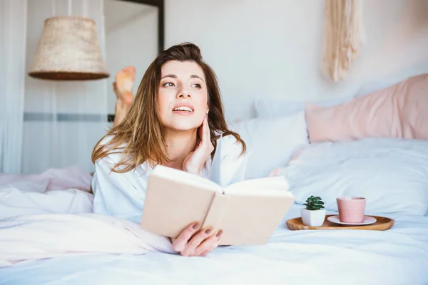 Menina lendo um livro, bebendo café ou chá, descansando na cama . — Fotografia de Stock