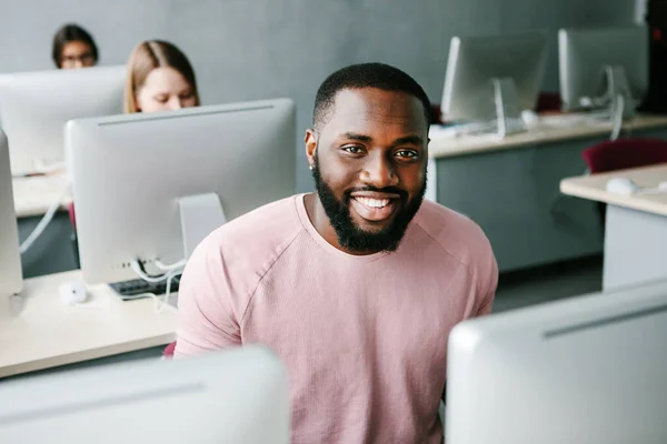 Vrolijke Afrikaanse man met behulp van de computer en glimlachen tijdens het zitten in het kantoor Rechtenvrije Stockfoto's