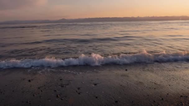 遠くの山と日の出の遅い動きでビーチに衝突する波 — ストック動画