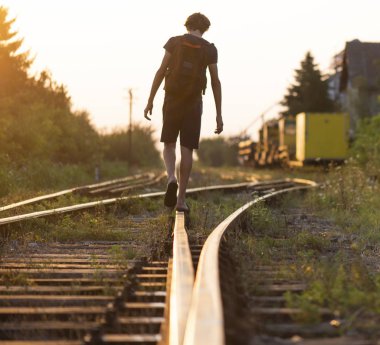 Elinde çanta olan yalnız bir çocuk raylarda yürüyor. 