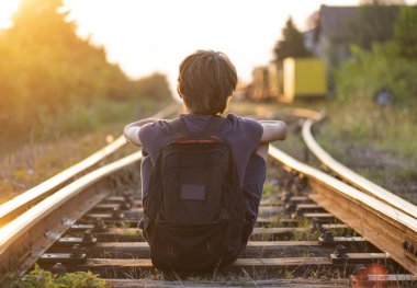 Elinde çanta olan yalnız bir çocuk raylarda oturur.