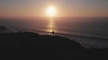 Gün batımında elleri okyanusun üzerinde uçurumun kenarında duran mutlu çocuk. Drone çocuğun üzerinden uçar ve okyanusun üzerinden günbatımına doğru uçar. Eski moda, kazanma kavramları, özgürlük, gençlik, mutluluk vs..