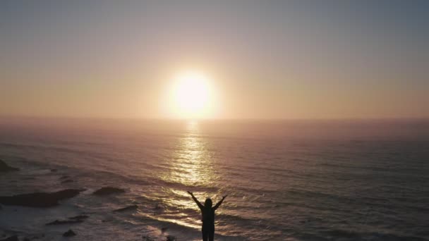 Donna felice con le mani in alto sulla scogliera sopra l'oceano al tramonto. Drone vola sopra la ragazza e vola verso il tramonto sull'oceano. mood vintage, concetti di vincitore, libertà, gioventù, felicità ecc . — Video Stock