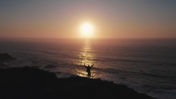 An optimistic birds eye view of a happy boy on cliff over ocean at sunset. The guy raises his hands up and rejoices in success, luck, victory, freedom, youth — Stock Video