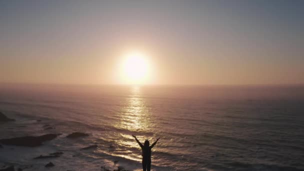 Mujer feliz con las manos en alto en el acantilado sobre el océano al atardecer. Drone vuela sobre la chica y vuela hacia el atardecer sobre el océano. Humor vintage, conceptos de ganador, libertad, juventud, felicidad, etc. . — Vídeos de Stock