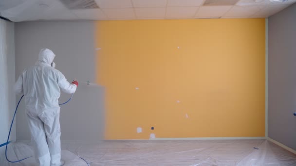 ホワイトの作業着を身に着けている労働者は、空気のないスプレー銃で白い色でオレンジの壁を塗装する。空飛ぶスプレー塗装。自宅やオフィスでの修理. — ストック動画