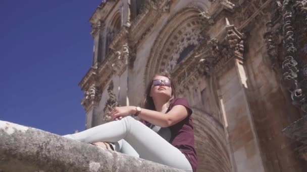 Kafkasyalı genç bir kız büyük antik bir binanın arka planında oturuyor, bir manastır. Arkasını döndü ve baktı. Turizm, gençlik, antik mimari konsepti. — Stok video