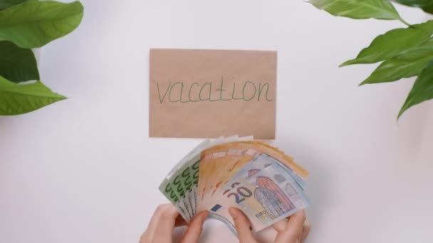 Bovenaanzicht. Vrouwelijke handen tellen Europees geld, euro, en zetten het dan op een envelop met een inscriptie, het woord Vacation. Achtergrond is een witte tafel met groene planten in de hoeken. — Stockvideo