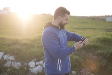Sakallı koşucu dışarıda antrenman yaptıktan sonra dinleniyor, telefonu akıllı saatle karşılaştırıyor.