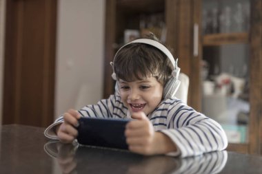 Evdeki çocuk dünyadan soyutlanmış kulaklıklarla telefona bakıyor.